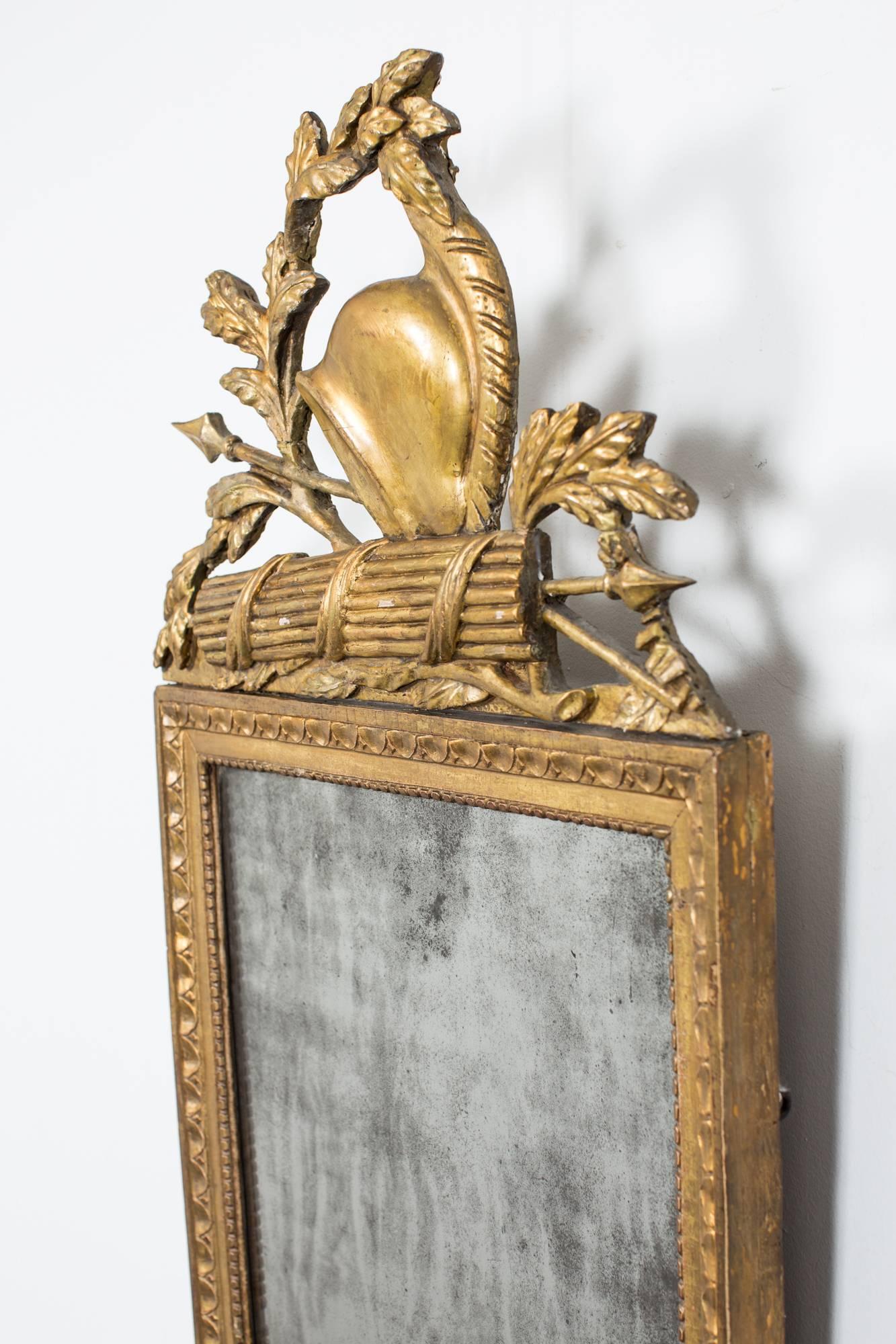 A wall mirror made during the 18th century in France. Frame in giltwood decorated with carved symbols such as a helmet, spier and leaves. Original glass.