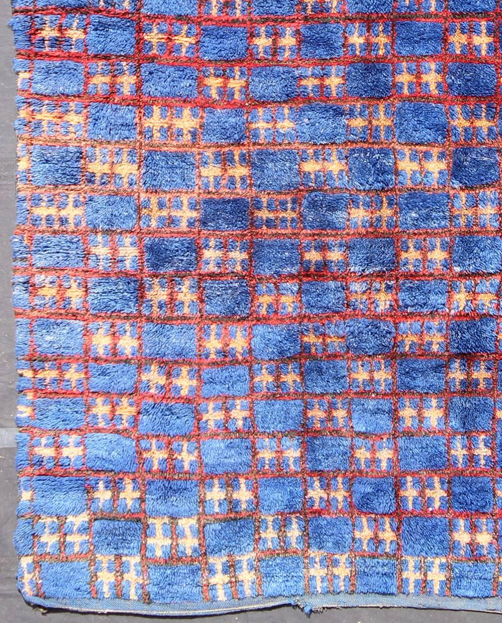 This unique vintage Moroccan carpet is characterized by a saturated, bold blue color and dramatic berber symbols. The alternating checkerboard pattern is outlined in red with tan cross motifs while its thick pile has a soft feel and a gorgeous aged