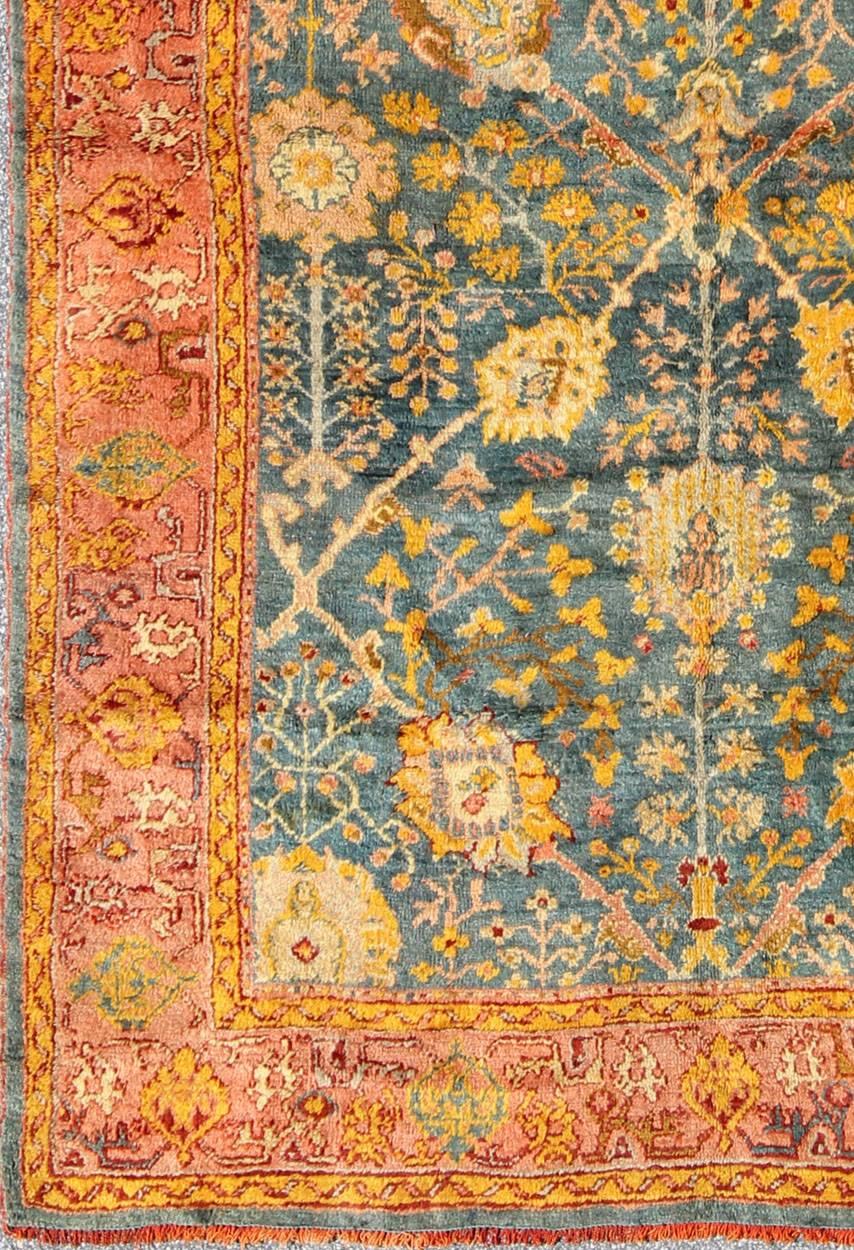 A coral/salmon border and a blue/blue-green background pleasantly dominate the entire body of this beautiful antique Oushak carpet. A spectrum of flowers, leaves, rosettes, tulips, peonies, lotuses and vine scrolls of all sizes gracefully float