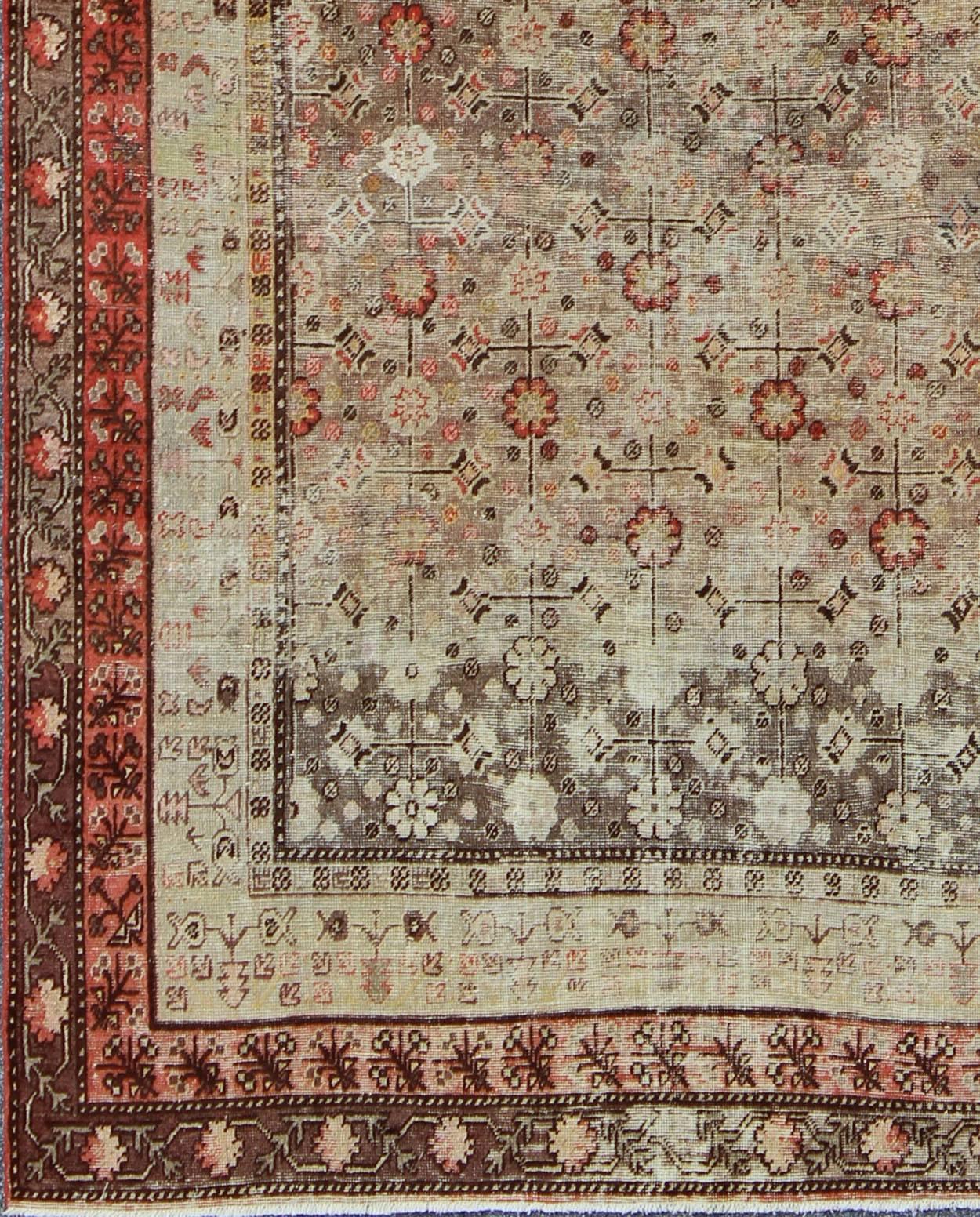 Measures: 6'3'' x 11'5''.
This Central Asian Khotan rug is an antique from the early 20th century. This piece features a traditional all-over flower pattern on a charcoal to tan field surrounded by multiple complementary floral borders of coral