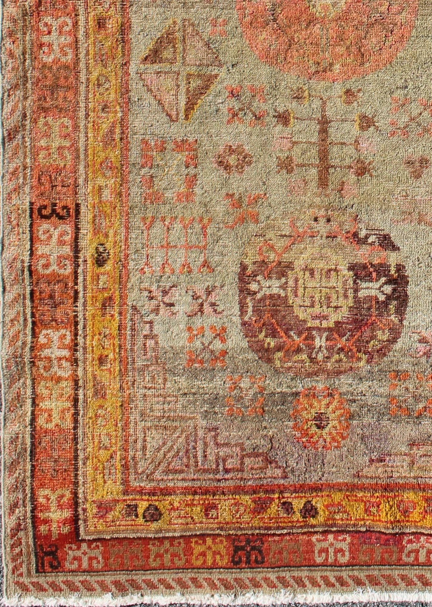 This antique, Central Asian Khotan rug from the early 20th century features a traditional, all-over floral, geometric pattern set on a silver-gray field. The central field is surrounded by multiple complementary floral borders of coral flowers with