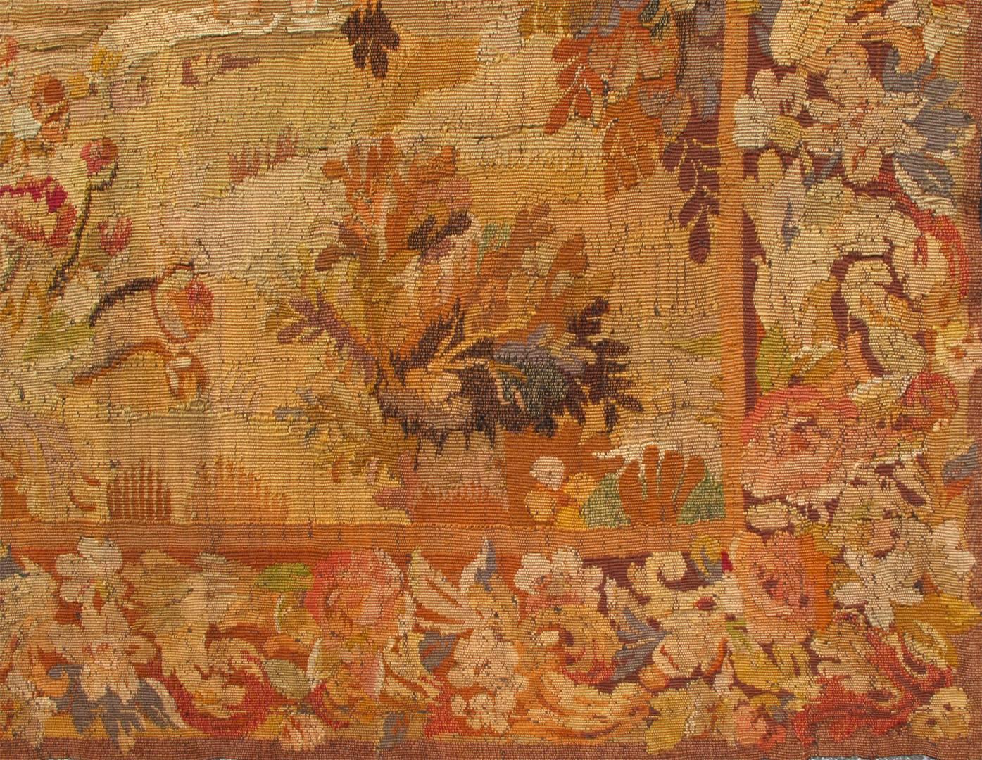 This engaging antique French tapestry depicts a traditional waterfront landscape framed by an expansive and richly decorative border of blooming flora. The entire scene is set on a waterfront surrounded by abundant trees, bushes and various flora.