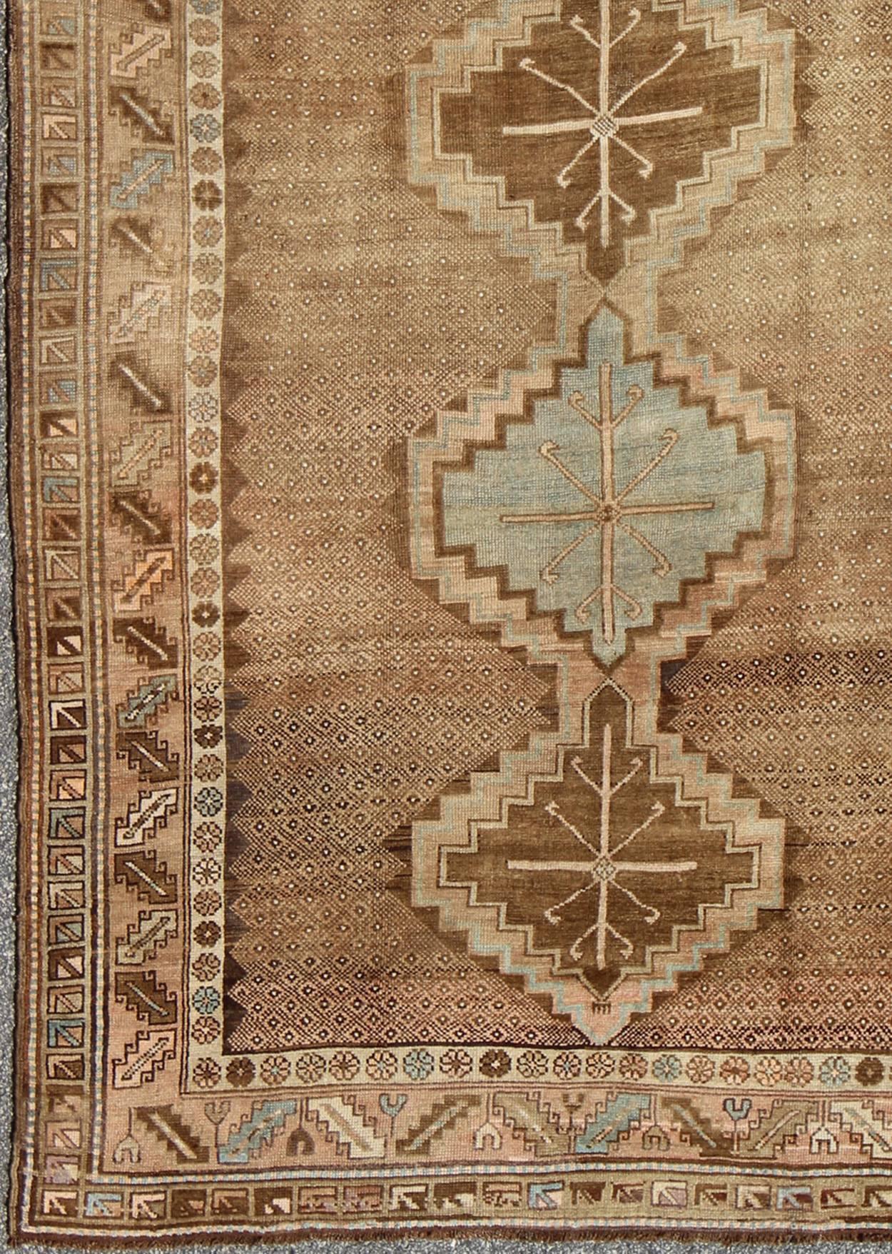 Set on a camel and brown background and accented by neutral cream and light blue, this tribal Turkish Oushak rug showcases five geometric medallions that stretch throughout the entire field of the rug. A multi-tiered border with repeating geometric