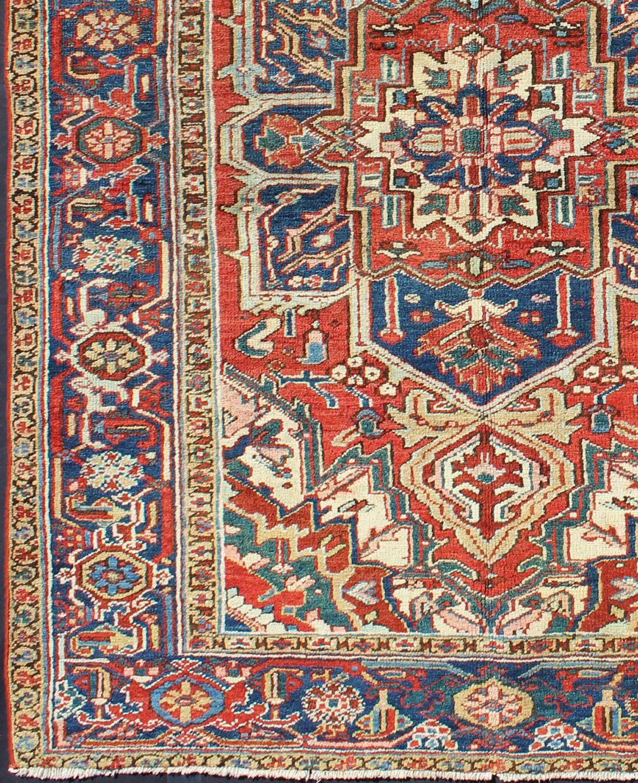 This antique Persian Heriz-Serapi from the early 20th century bears an exquisite design rendered in gorgeous, warm hues of red and denim blue. A highly stylized geometric center medallion anchors the piece and is surrounded by numerous geometric