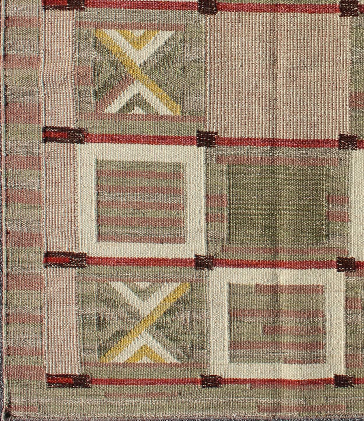 This Scandinavian flat-weave patterned rug is inspired by the work of Swedish textile designers of the early to mid-20th century. Featuring a unique blend of historical and modern design, rendered in shades of green, light mauve, red, brown and