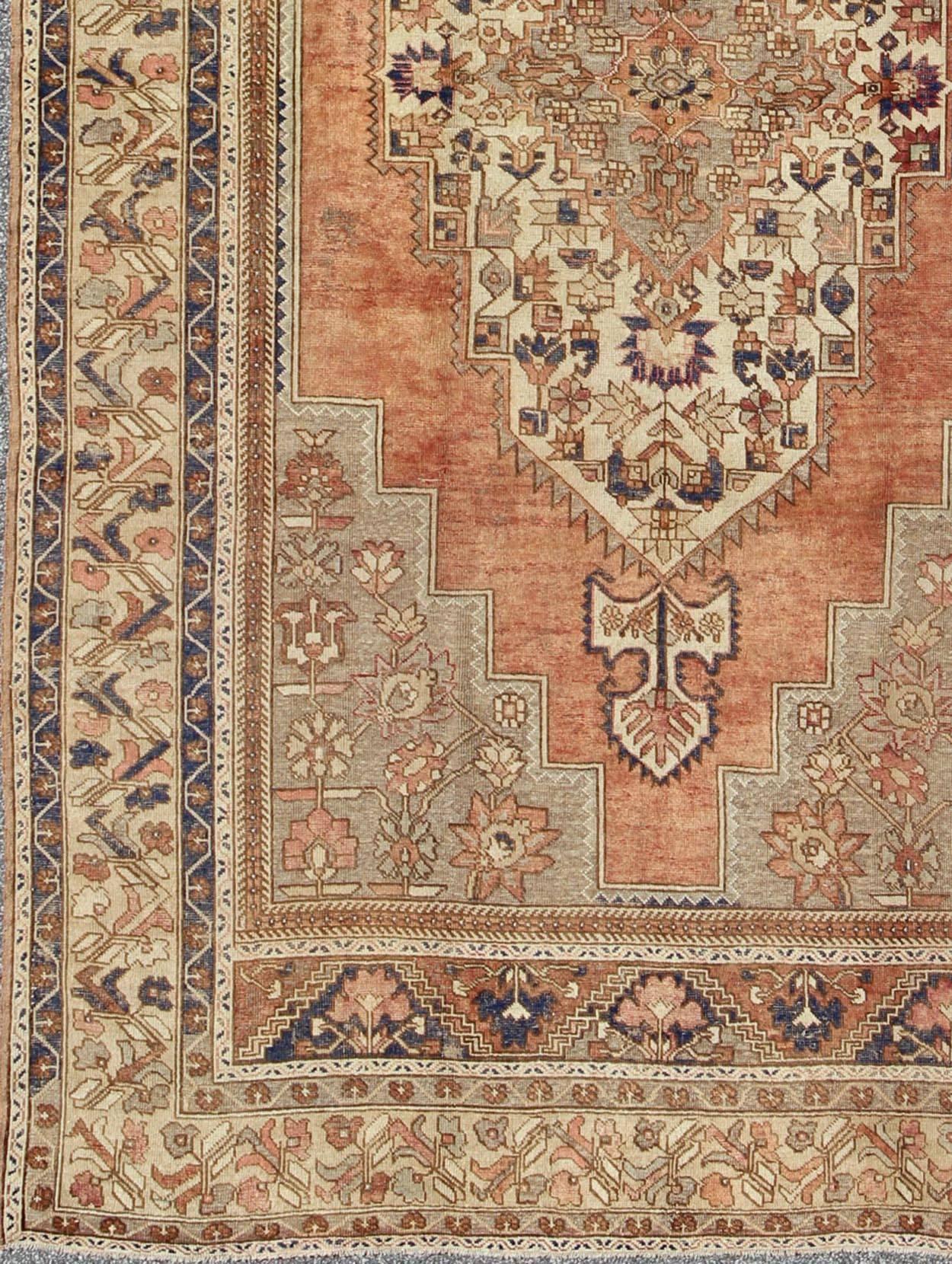 This beautiful Turkish Oushak Rug has a medium blue color with a light orange background. The center medallion has earth tones with blues. The border has a rolling flower pattern. There are colors such as creamy yellow, light green, grey blue,