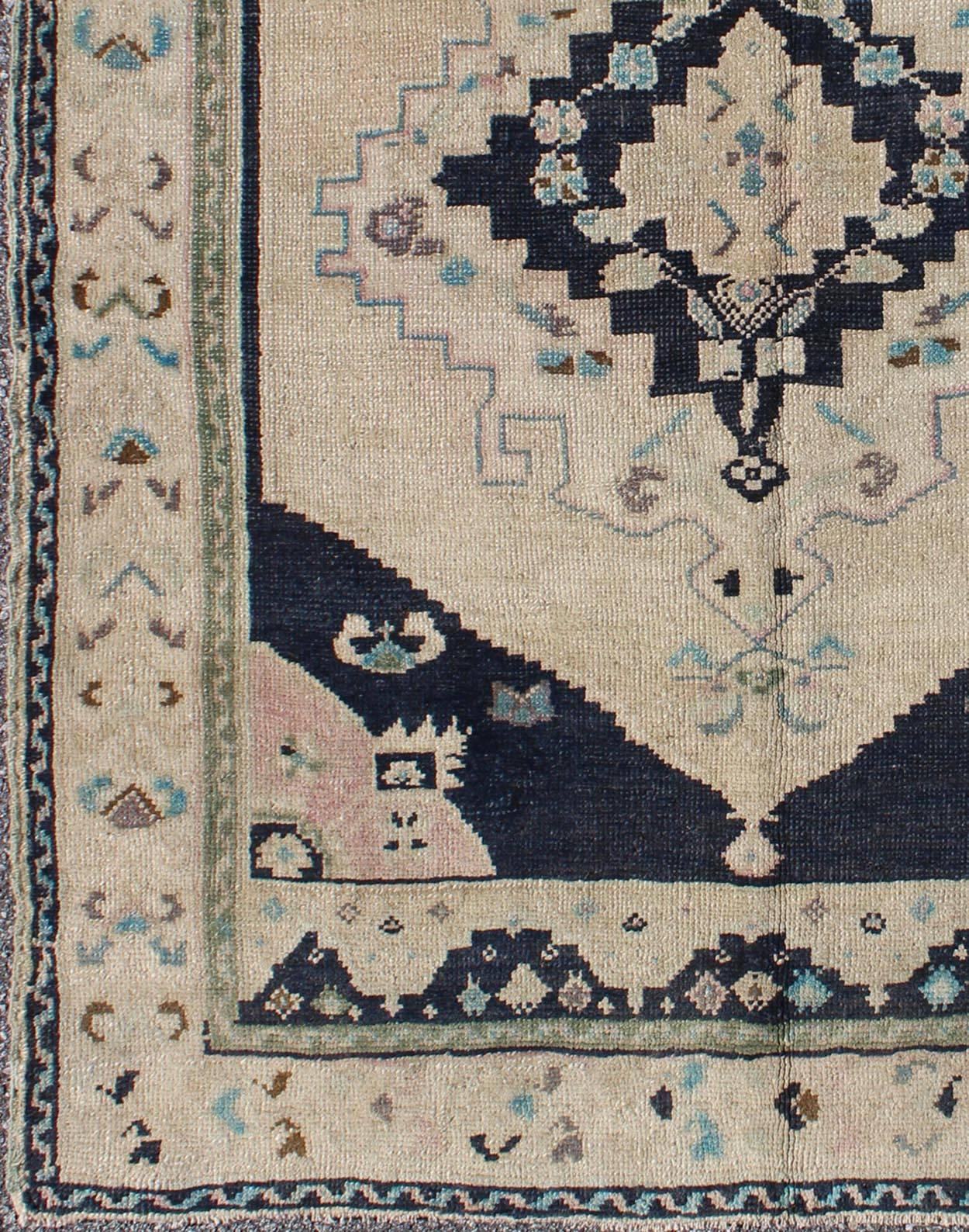 This beautiful rug from mid-20th century Turkey features a medallion with floral motifs surrounding it. The cream ground is home to the center medallion. Four dark blue cornices hold the field with hints of pink. A soft border with floral and leaf