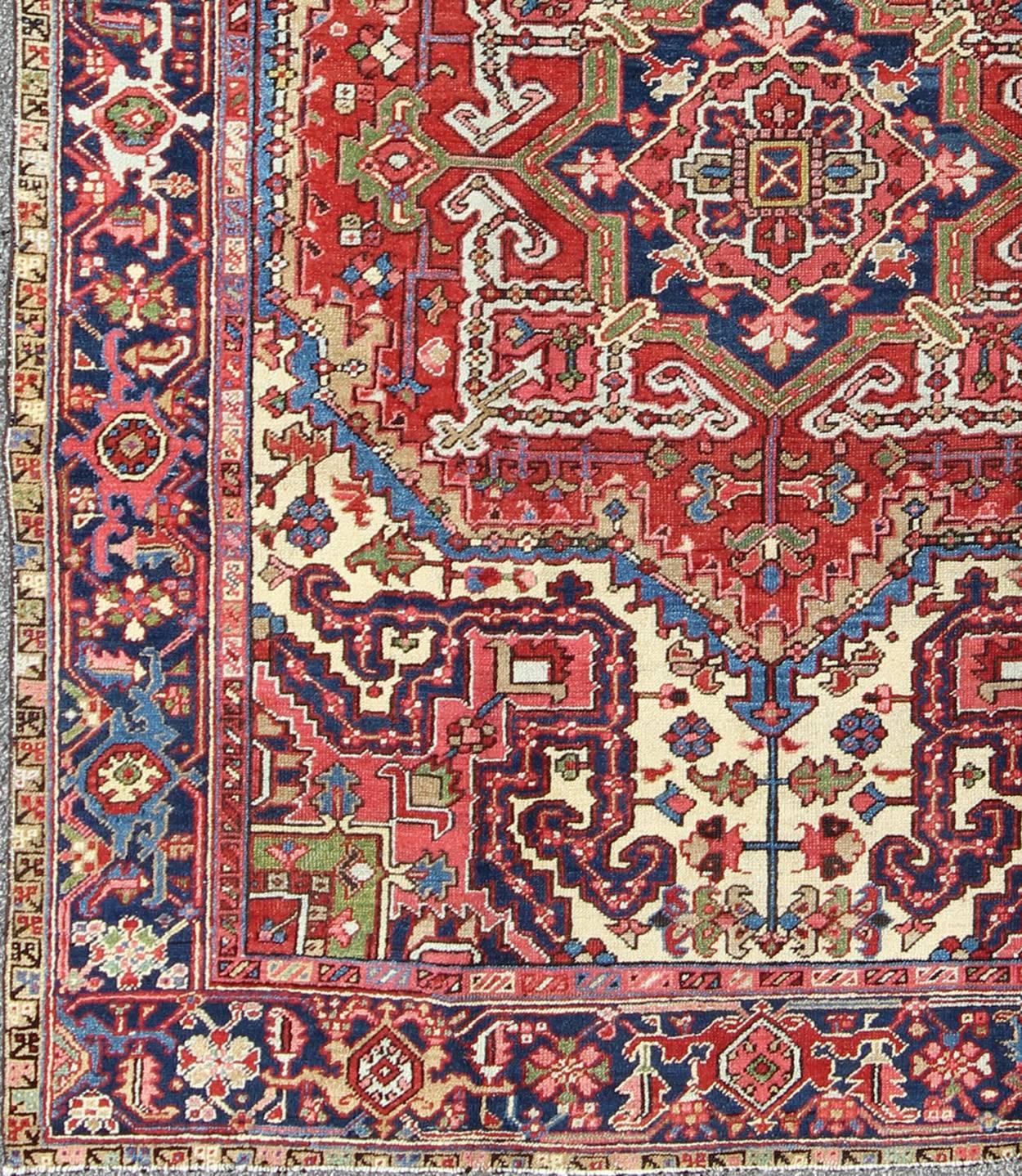This magnificent antique Persian Heriz carpet from the early 20th century bears an exquisite design rendered in gorgeous, warm hues of red and various shades of blue. A highly stylized geometric center medallion anchors the rug and is surrounded by