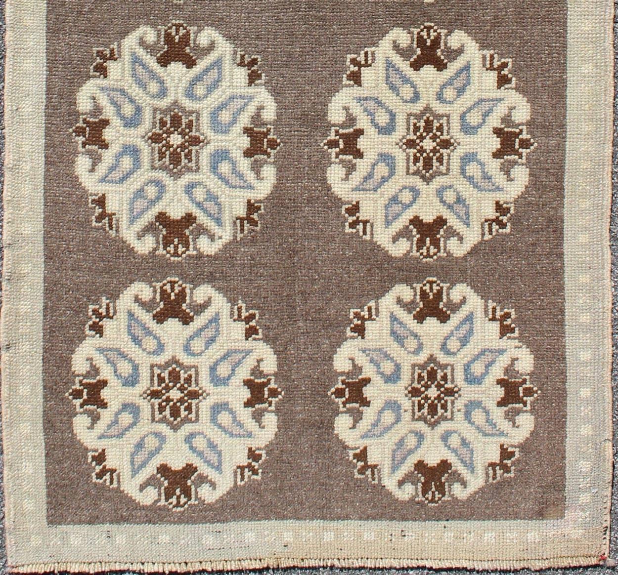 This Turkish Oushak gallery rug features ten floral medallions that take center stage on a brown field. The border reveals tribal inspiration in soft shades of taupe, cream, blue, and chocolate brown accents. 

Measures: 2'9'' x 6'4''.

Tribal