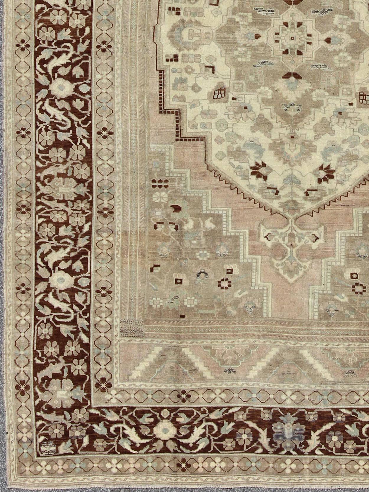 Measures: 7'5 x 11'4.
This rug showcases a large central medallion surrounded by a gentle pink color. A cream medallion is filled with floral designs of pale blue, light green, and shades of neutral browns. The chocolate brown outlines in the