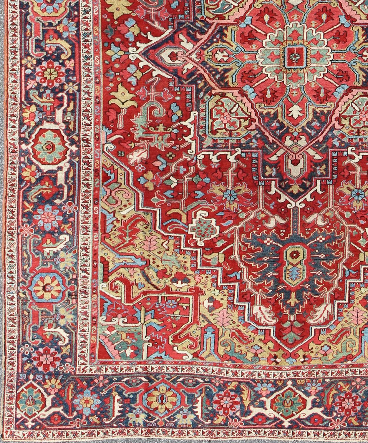Dieser prächtige antike persische Heriz-Teppich aus dem frühen 20. Jahrhundert zeigt ein exquisites Muster in wunderschönen, warmen Rottönen und verschiedenen Blautönen. Ein stark stilisiertes geometrisches Medaillon in der Mitte des Teppichs wird
