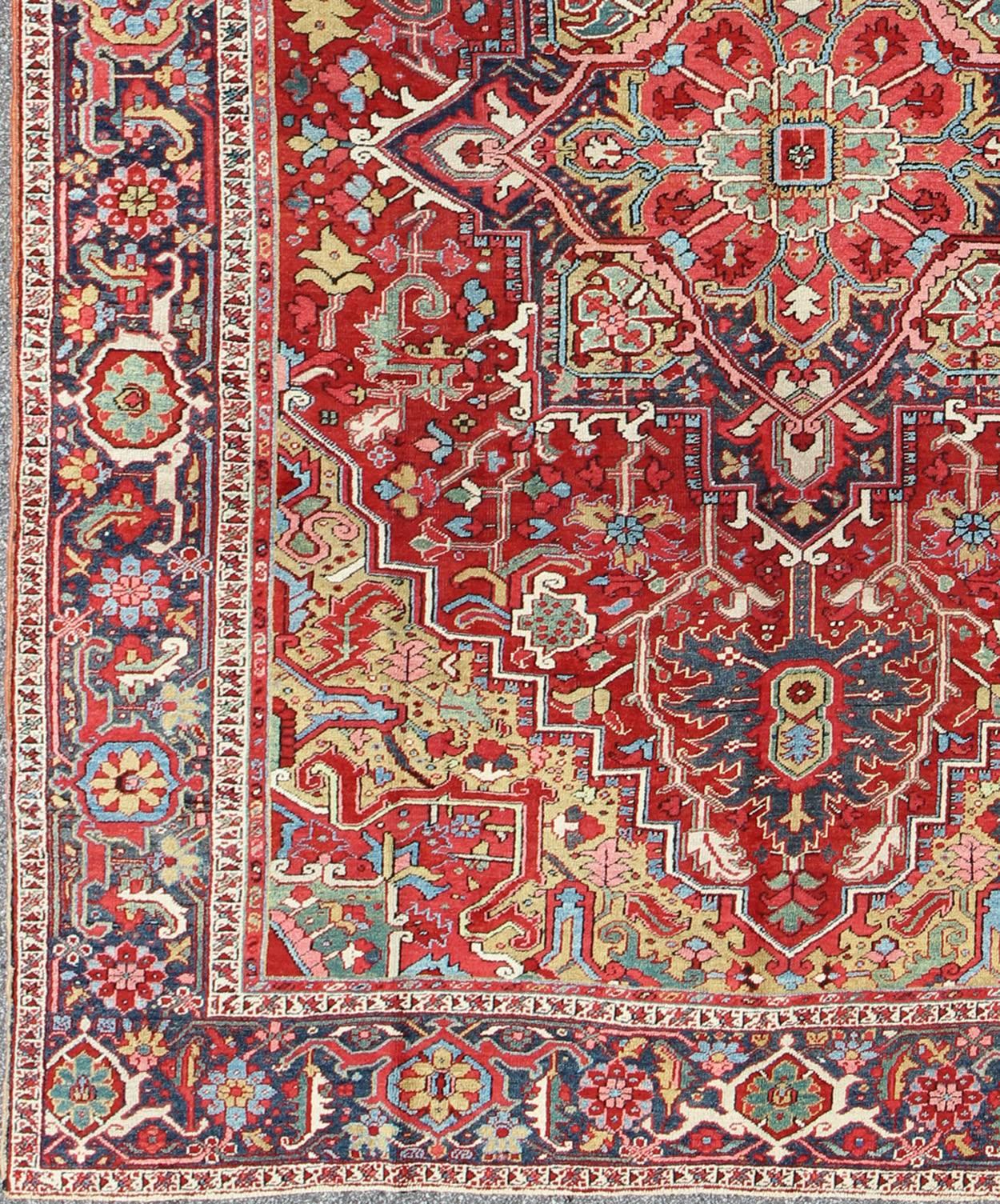 This magnificent antique Persian Heriz carpet from the early 20th century bears an exquisite design rendered in gorgeous, warm hues of red and various shades of blue. A highly stylized geometric center medallion anchors the rug and is surrounded by