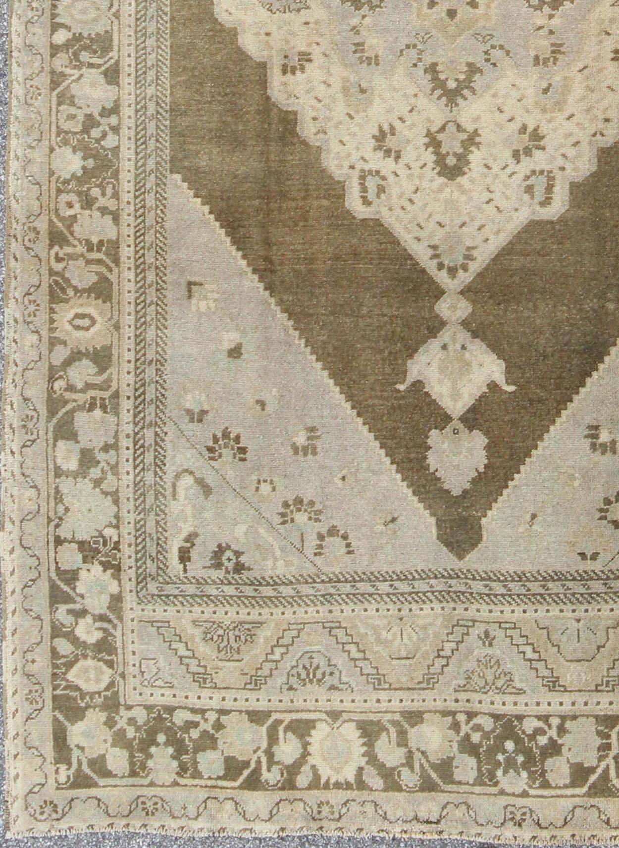Dieser auffällige türkische Oushak-Teppich im Vintage-Stil hat einen braunen Grundton, der durch ein schillerndes Medaillon-Muster in verschiedenen Farbtönen hervorgehoben wird. Ein großformatiges Medaillon erstreckt sich über das mittlere Feld. Die