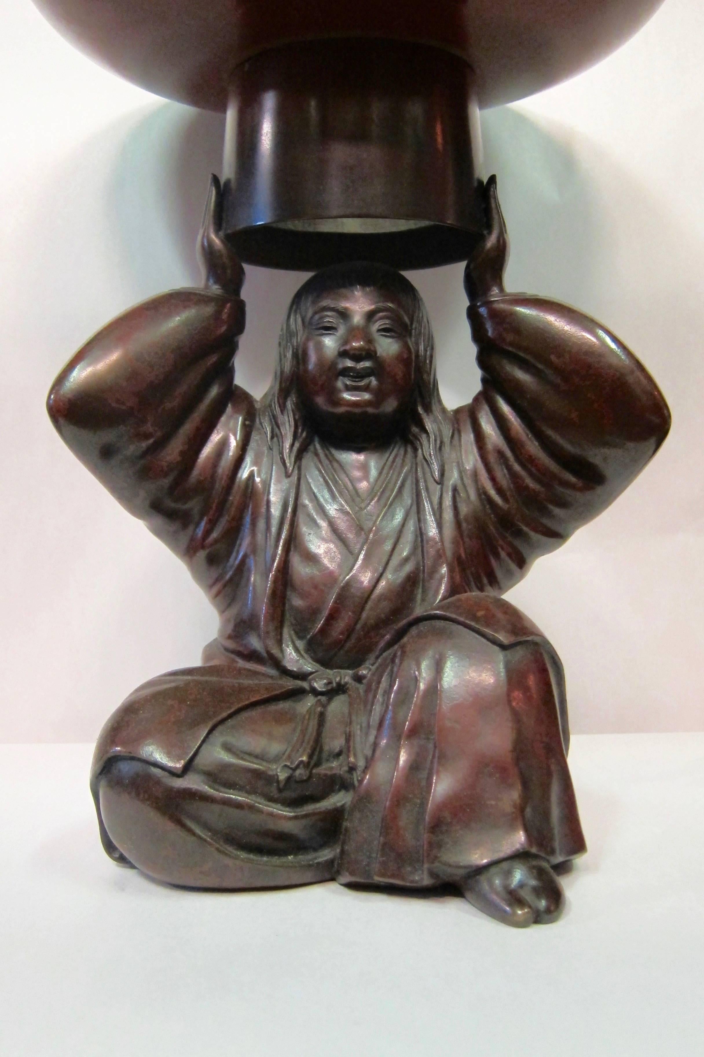 This beautifully sculpted 19th century detailed bronze sculpture features a robed Japanese man supporting a wide hollow circular form above his head. A wide bronze form inserts into this opening and may hold a variety of things including a plant,