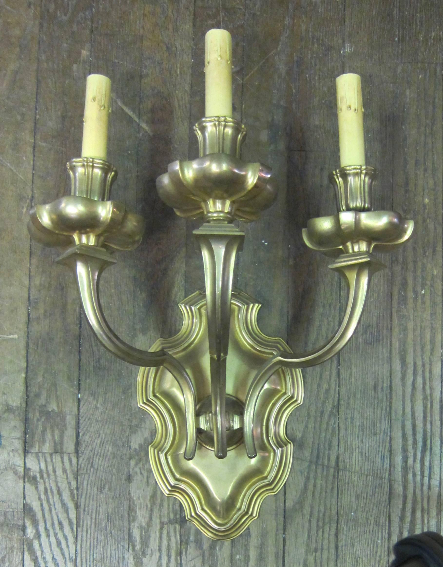 This vintage pair of gold plated bronze wall sconces was designed & produced in the 1920s by E F Caldwell. The sconces have a stylized shield backplate with a rippled rim. Three dramatic arms that flare upward & terminate into candleholders
