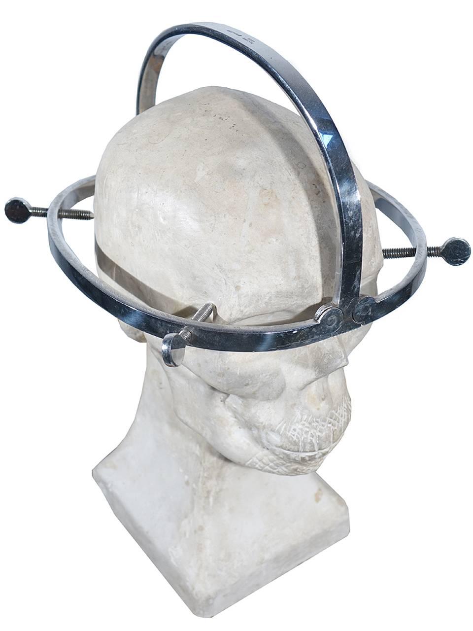 These devices were used by surgeons as well as morticians. This example is hinged and the sides fold up next to the top bow. The plaster skull is also included to display the device. This 1920s skull was used as a teaching aid for medical students