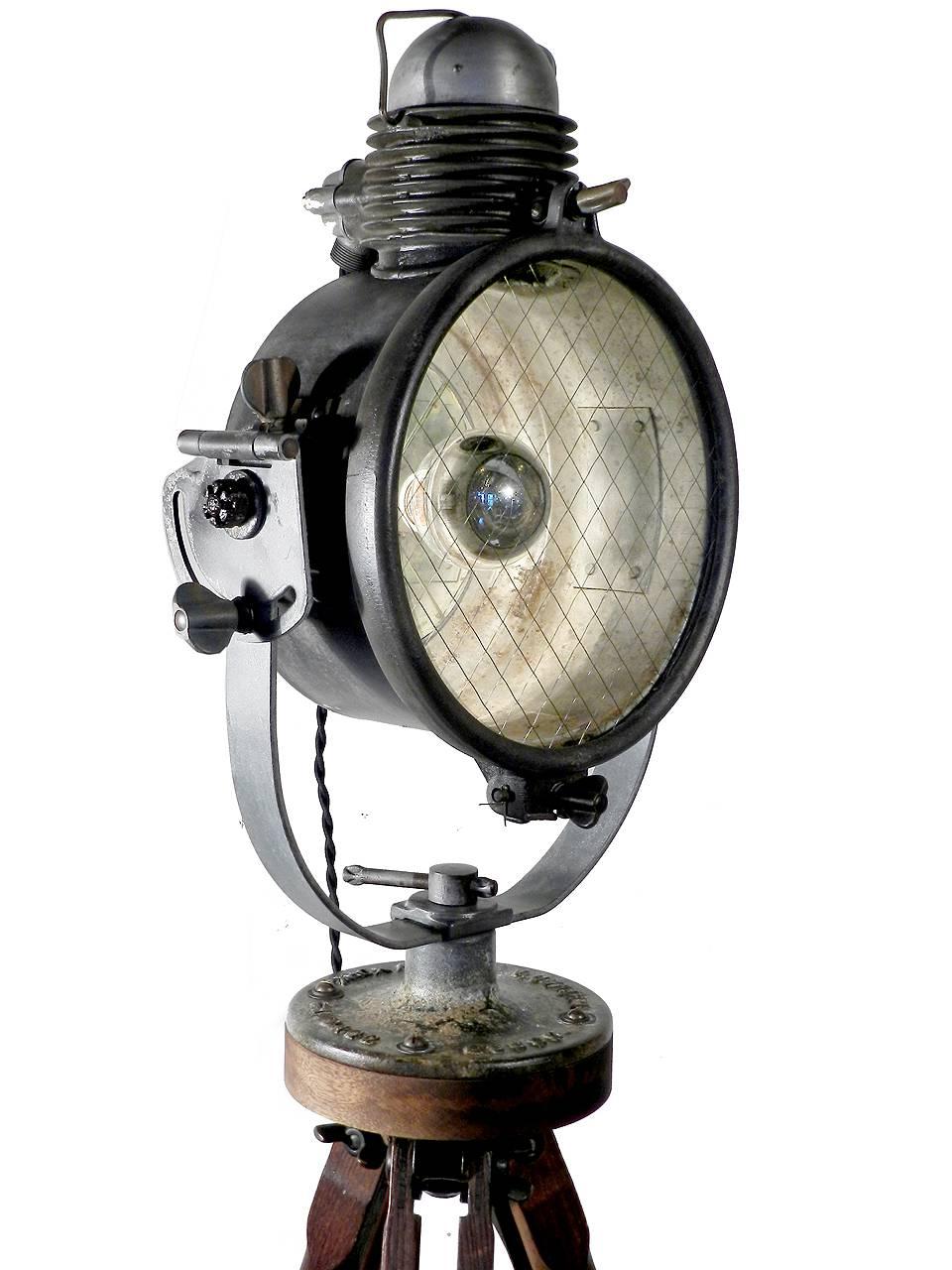 This is a very early flood light that has been rewired for home use and standard bulbs. It has a wonderful Industrial look with a wire glass lens, mirrored reflector, adjustable wood tripod and black twisted fabric covered wire. The iron fixture has