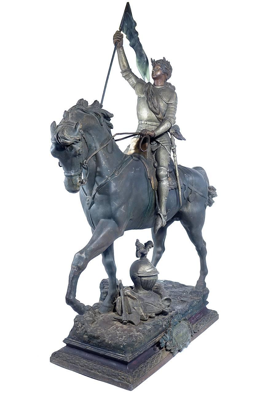 This is a large antique French shelter sculpture depicting Joan of Arc. She is riding on a horse in full armor carrying a flag. In addition Her helmet, ax, shield and gloves are depicted on the ground. The plaque on the front base reads. JEANNE dArc