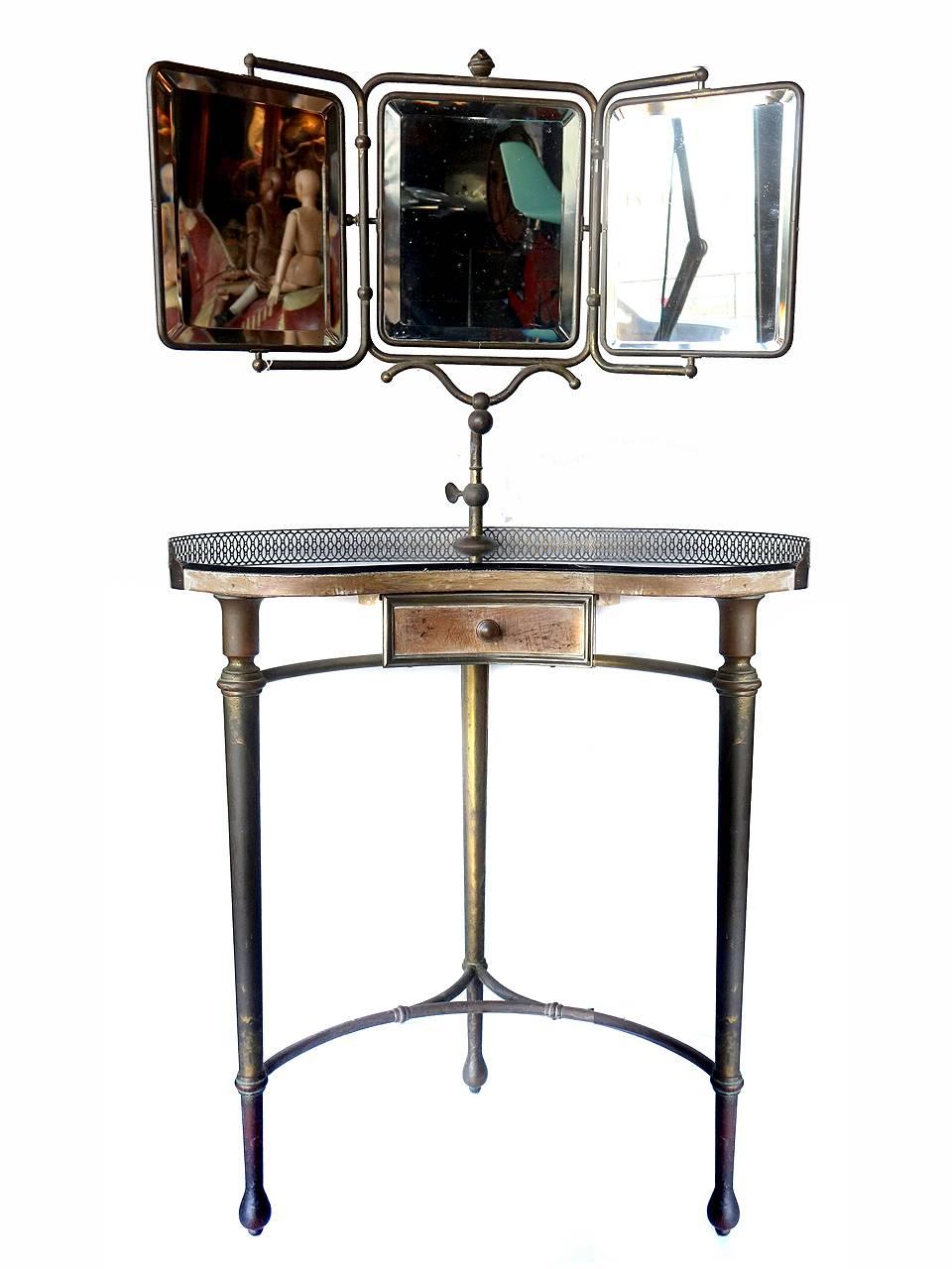 This early original table has a black glass top a d three beveled mirrors. The mirror frames and legs are brass having and dark aged patina. The table and draw are wood. I think the tables edge may have had a fabric fringe tacked on the front edge.