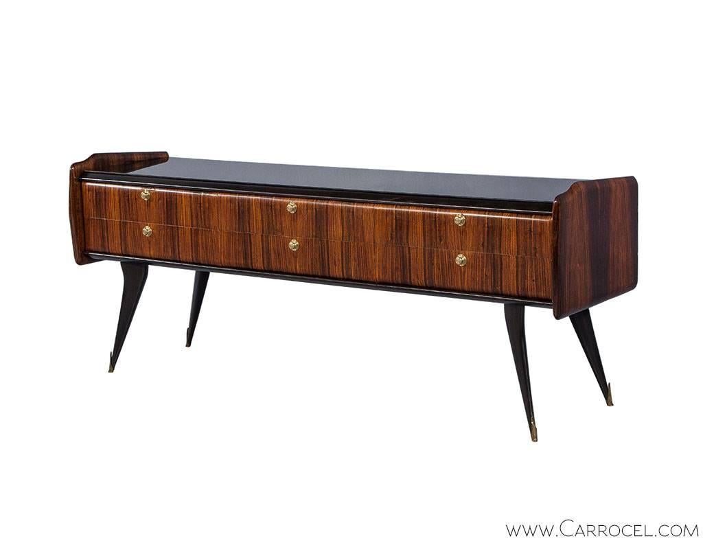Gorgeous rosewood credenza with a long linear shape contrasted by subtly filleted edges. The piece has ben designed with a black glass top, a veneered facade divided into six drawers with small brass pulls original to the piece and black diagonally