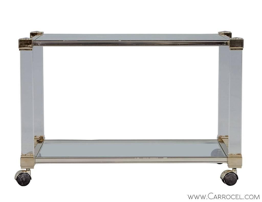 Delicate and light, this Lucite and brass side table is designed in the manner of Pierre Vandel. With a tubular design, Lucite legs extend between an upper and lower brass frame supporting a glass top and lower shelf. The table has a multifunctional