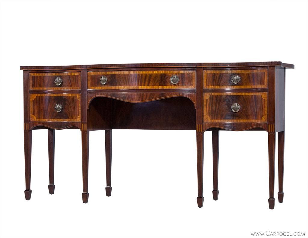 This sideboard by Berkey & Gay Furniture has a flamed mahogany design with contrasting satinwood borders. The serpentine style lends to the Hepplewhite heritage and the brass pulls add texture and beauty. The two top side drawers have dividers for