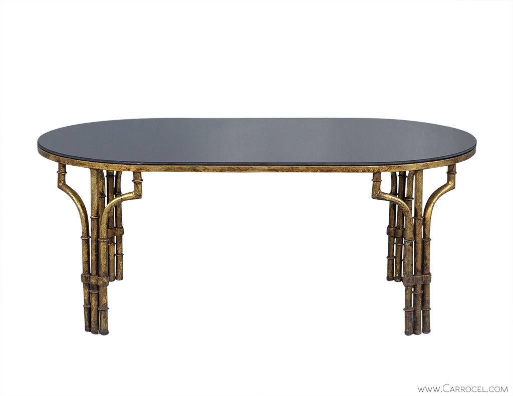 This unique cocktail table is the epitome of contrasting style. The brass base boasts a distressed gold finish and is topped with oval shaped, black glass. The smooth texture of the glass compliments the rough, bamboo style table legs to truly