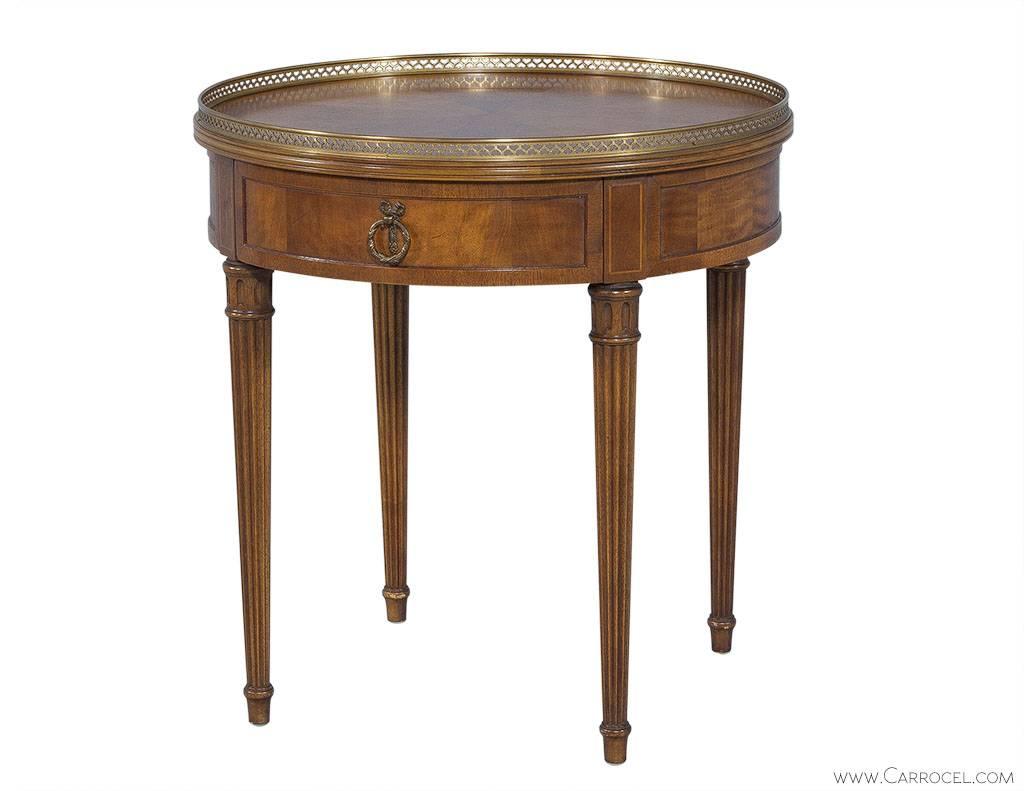 This rounded walnut side table is adorned with ornate brass accents. It sits atop reeded, tapered legs and is full of detail and timeless flair. The veneered top showcases a starburst design and brass gable around the edge. The single drawer is