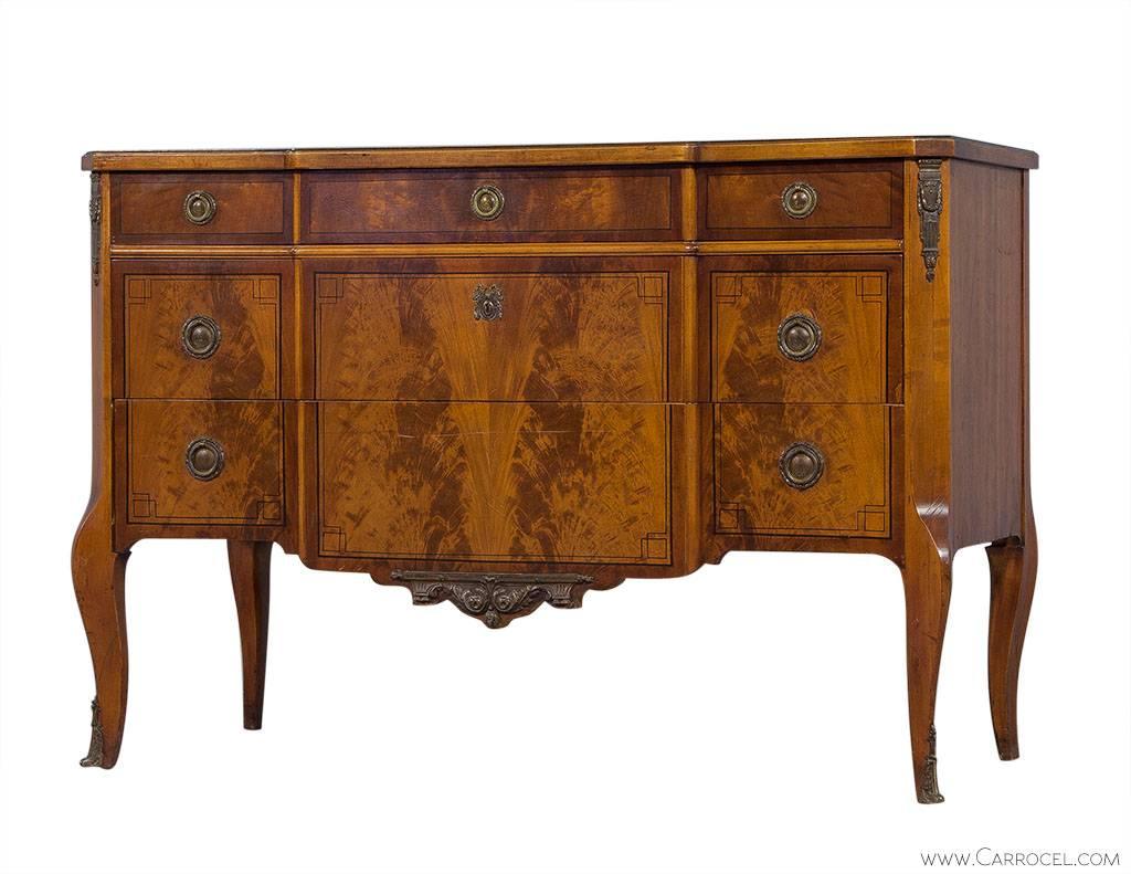 This Louis XV style commode is made of mahogany with a lightly distressed finish. The original laurel wreath brass ring pulls, along with the brass mounts on the legs and façade and decorative escutcheon, epitomize the character of this piece. This