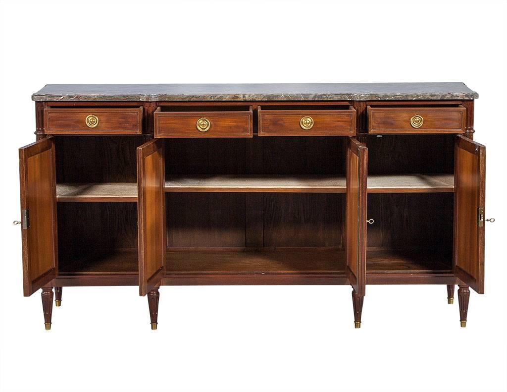 This gorgeous Louis XVI style buffet is a timeless addition to any dining room. It is made of stunning mahogany and is framed by fluted columns that end in brass-capped legs. This piece is topped with grey veined marble, adding a bit of smooth