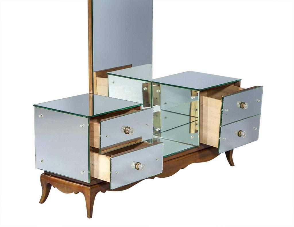 An absolutely stunning piece, this mirrored vanity carries a luminous and grand essence of days gone by. With a touch of deco influence and a Hollywood Regency appeal, the vanity is enveloped in mirror with left and right double-drawer banks,