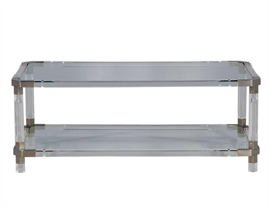 This French style cocktail table is quite sharp. It showcases brushed metal corner connectors with acrylic stretcher bars and a glass top and lower shelf. The corners are made of steel but angled to soften the edges, as the style remains sleek. A