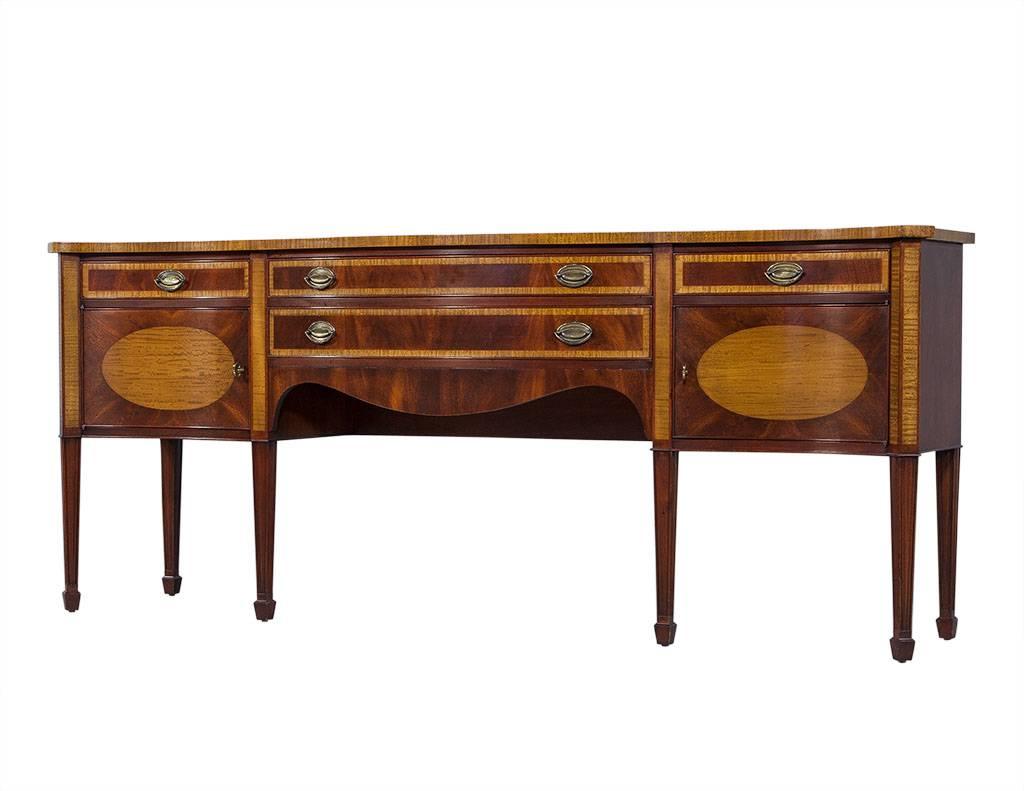 This Louis XVI style sideboard is crafted out of solid mahogany. The case is carved into a serpentine shape, and it has satinwood accents and inlay. The case also houses two cabinets and four drawers that sit atop fluted, tapered legs. Adorned with