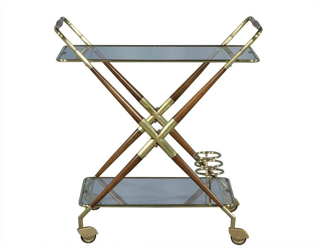 This Mid-Century Modern bar cart is absolutely striking. This mixed media piece is crafted out of alternating wood and polished brass to form an X-frame on four wheels. There are two glass shelves with a bottle stand on the lower tray as well, and