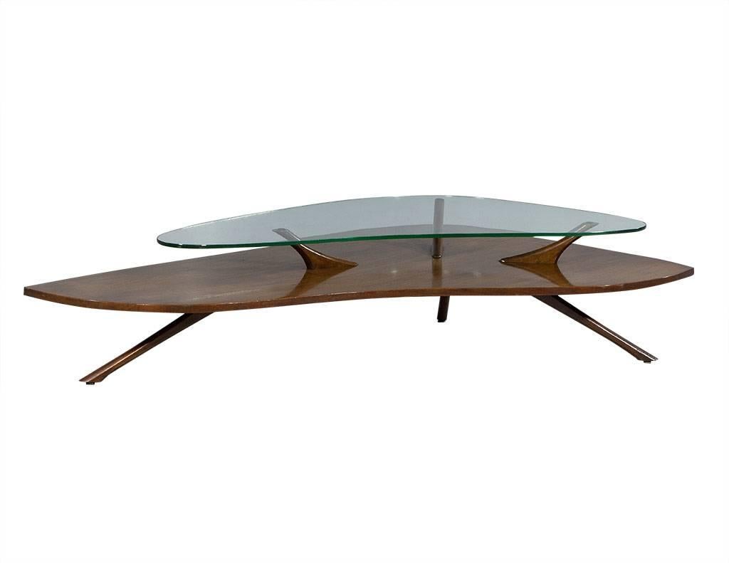 This Mid-Century Modern cocktail table is futuristic and stylish. The wood base is carved into a kidney shape and finished in light walnut with a glass top. It sits atop three angled legs on bottom and three legs between the wood and glass top. A