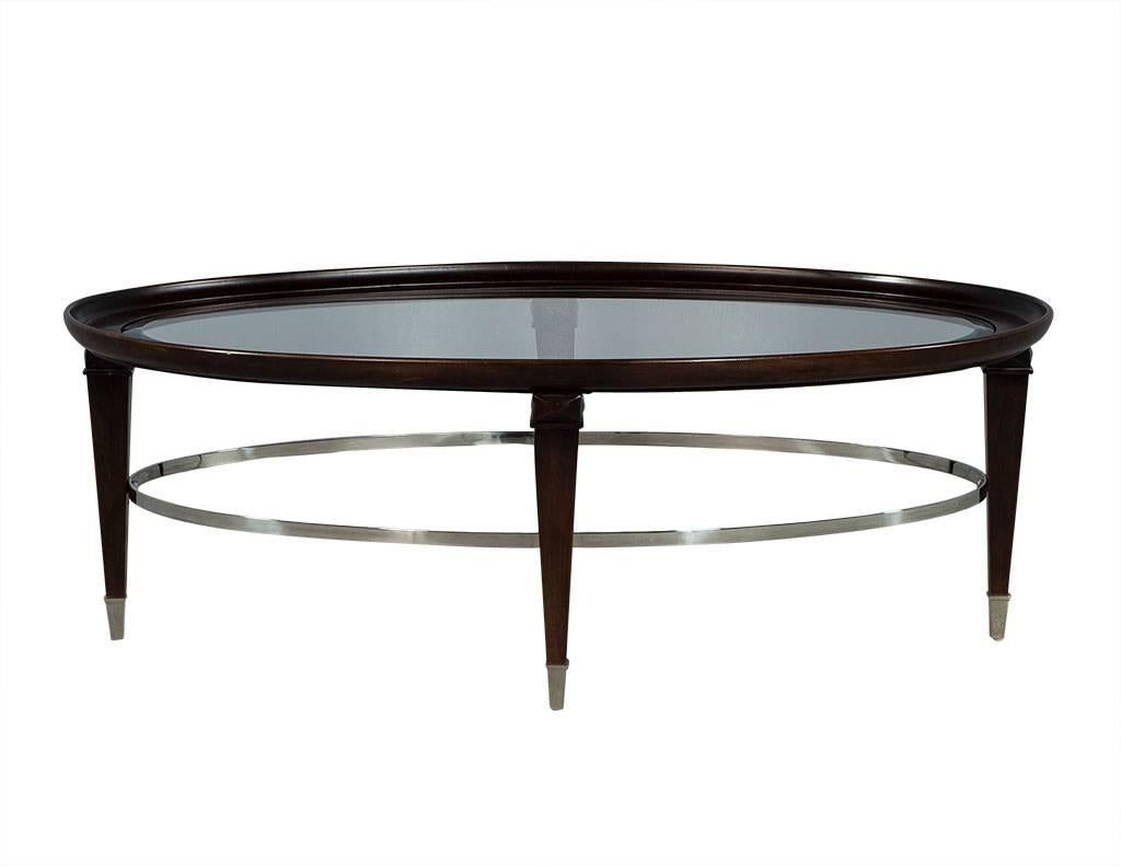 This transitional style cocktail table is absolutely stunning. It has an oval-framed mahogany base with an inset bevelled glass top sitting atop four legs with a polished nickel banding on the base and caps on the feet. A perfect fit for a richly