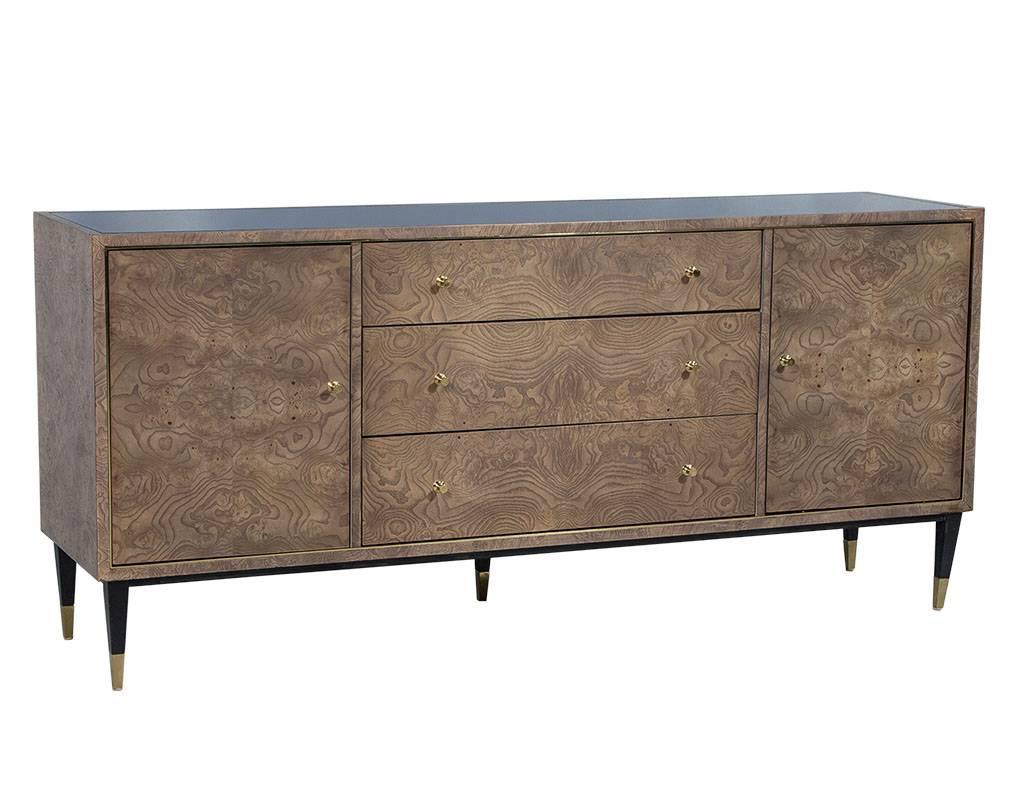 This contemporary credenza is made of burled wood with a black painted glass top and two end cabinets with three shelves and three center drawers. The piece sits atop fluted legs with polished brash feet and matching hardware. A fresh addition to