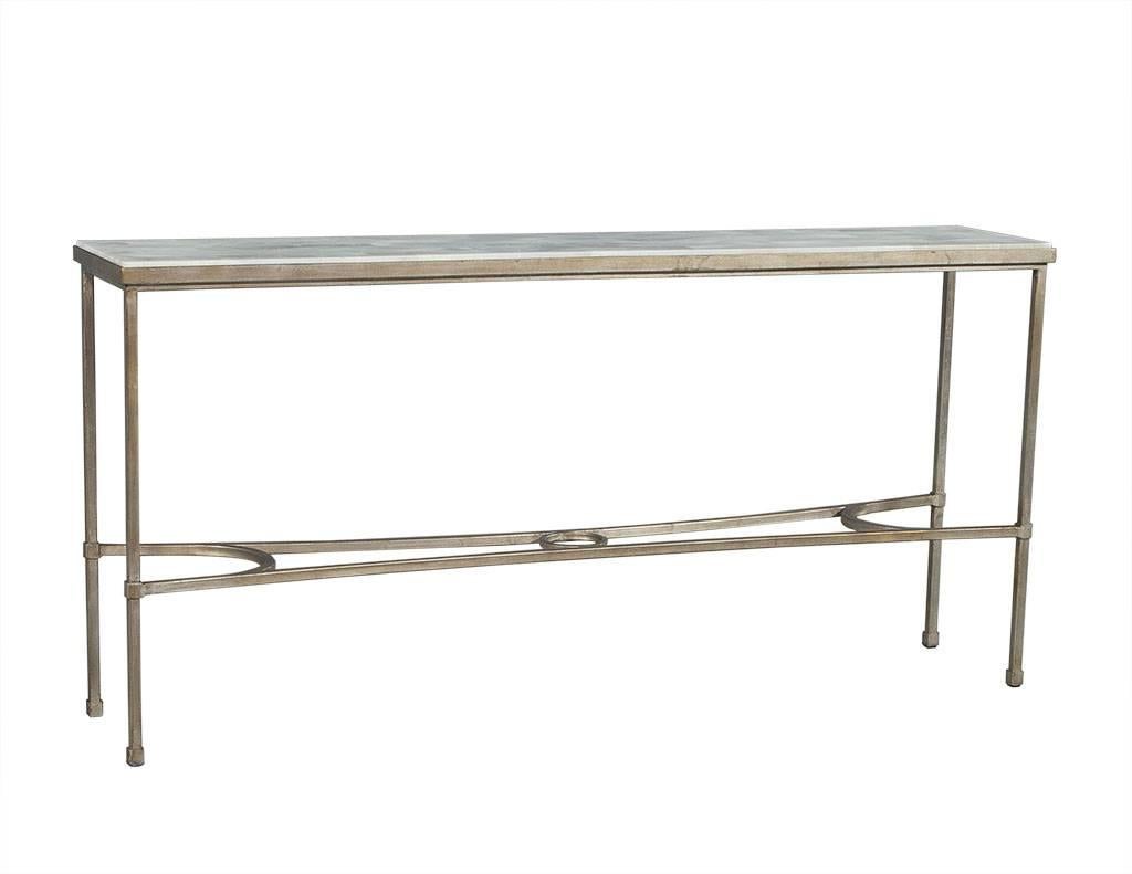 This transitional style console table showcases a split face, interlocking pattern white stone top inset on a rustic, four leg steel base in an aged gold finish. Connecting the legs is a demilune stretcher accented with a decorative center circle. A