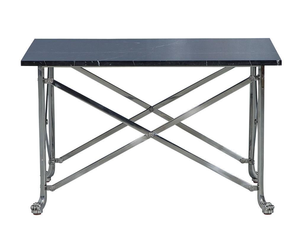 Cette table d'appoint néoclassique est une pièce très chic, avec un plateau en pierre noire du Burkina qui repose sur un cadre en acier inoxydable poli à croisillons. Les pieds en forme de pattes de lion ajoutent une touche dramatique, et font de