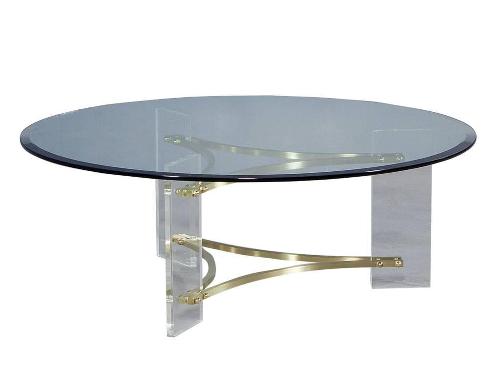 This elegant, Hollywood Regency style round cocktail table features a bevelled edge glass top and is supported by three Lucite panels, connected by slim demilune brass stretchers. Stunning addition to any sitting area!