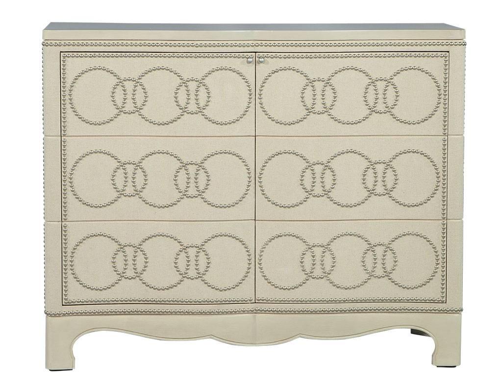 This transitional style cabinet is a beautifully designed piece, with white linen wrapped delicately around the cabinet doors and sides. Nailheads adorn the trim in a circular pattern, while the serpentine wood on top and scalloped base add the