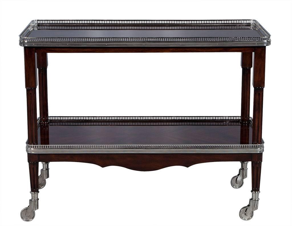 This exquisite two-tier bar cart is made for entertaining! A flamed mahogany frame is supported by four stainless steel castors and chinoiserie style banding on both lower and upper shelf parameters. Finished in deep walnut that adds a luxurious