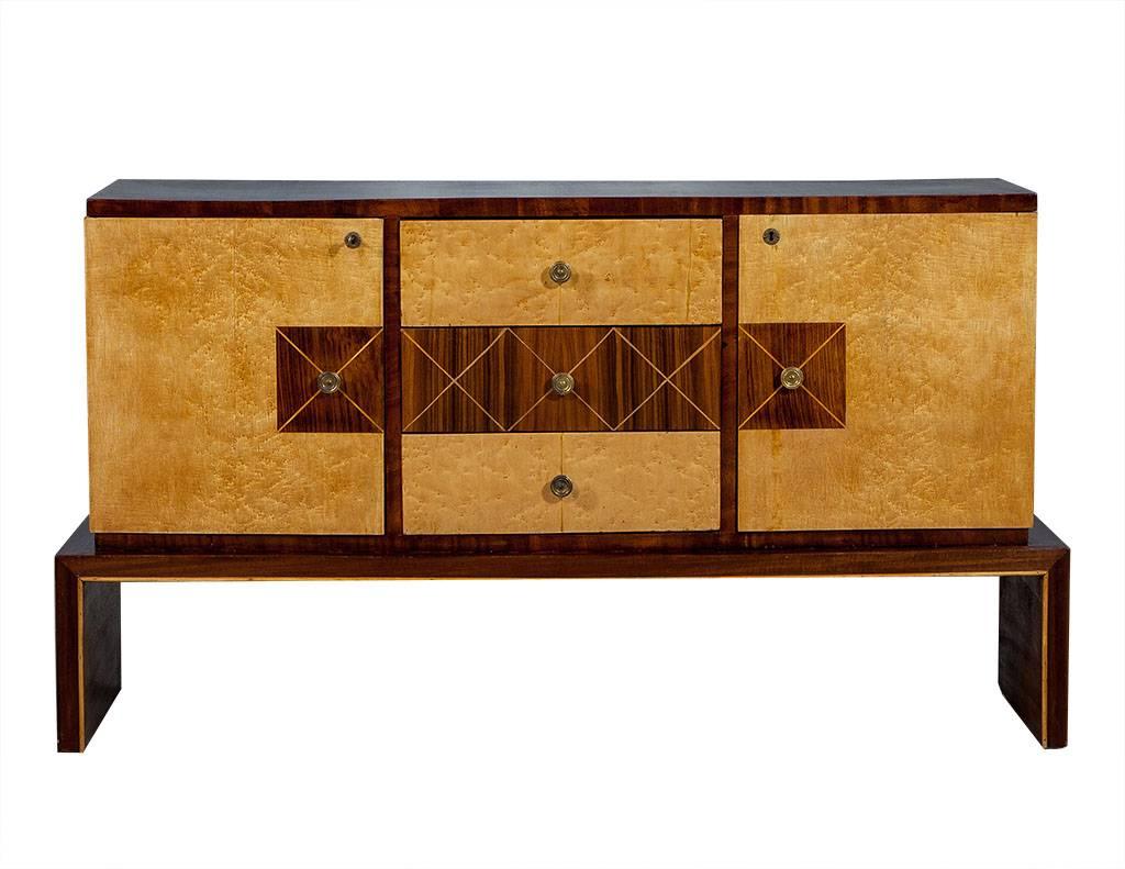 This Art Deco style buffet is crafted out of a rosewood frame with a raised waterfall base and left and right doors that conceal two shelves. There are also top and bottom central drawers with a veneer made of bird’s eye wood. The center panel is