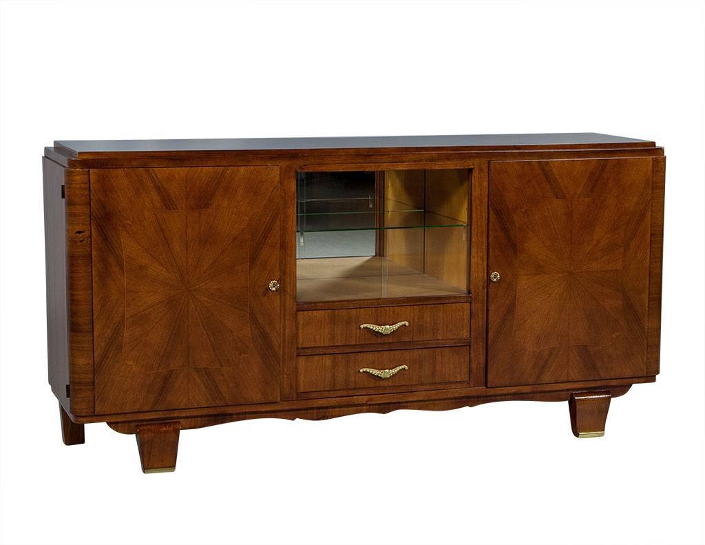 This Art Deco style buffet is crafted out of walnut with a starburst field on the left and right cabinet doors which conceal two shelves. The top is raised and there are rounded corners on the frame. The center compartment includes an upper display