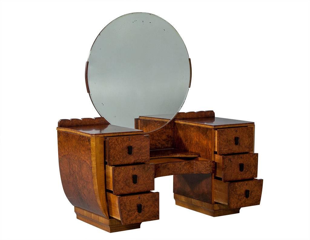 This Art Deco style vanity is crafted out of burled wood and finished in a medium walnut stain. It has a single drawer beneath a large, round antique beveled edge mirror surrounded by scalloped trim on both ends. The left and right compartments