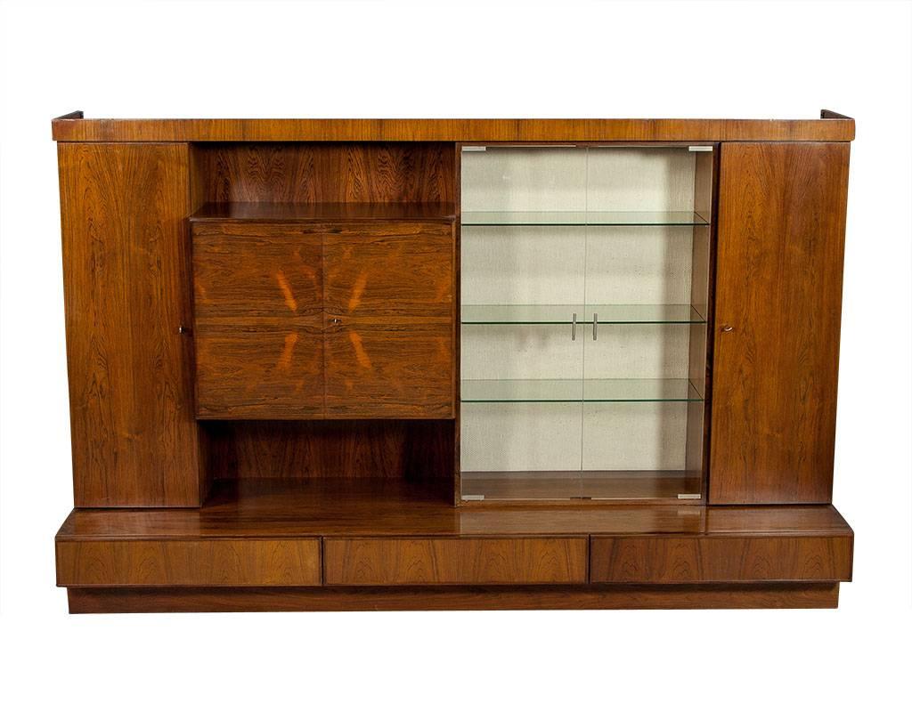 This Mid-Century Modern wall unit is beautiful and functional. It is crafted out of rosewood and finished in a medium walnut stain. There are three bottom drawers and doors at each side. The right side encases four wood shelves while the left