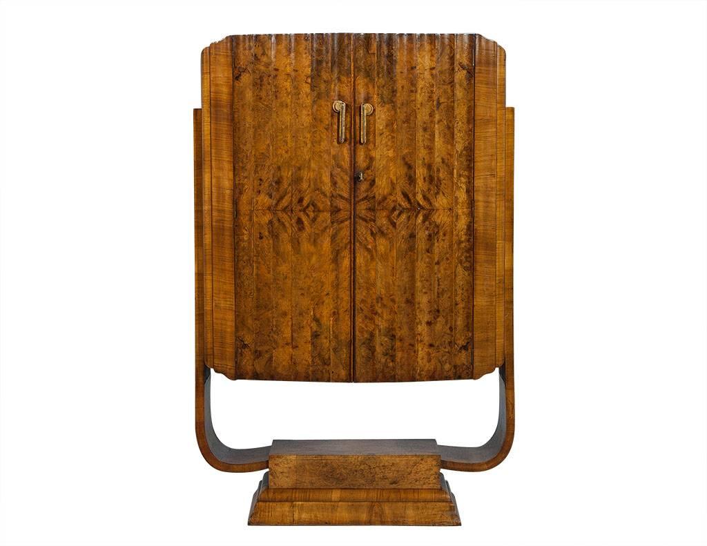 This Art Deco style bar is retro and unique. The U-shaped, floating cabinet is composed of blonde, mapa burl and walnut. It rests upon a raised plinth pedestal. Louvered double doors conceal the interior bar that is divided into two compartments.