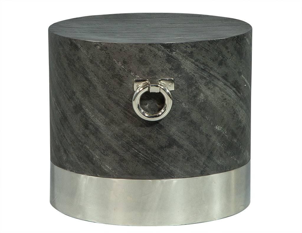 This modern end table is daring and unique. Composed of a thick, cylindrical, brushed stainless steel base accented with rings on either side and feature a grey, stone wrapped top making it modern and functional. A perfect fit for a boldly designed