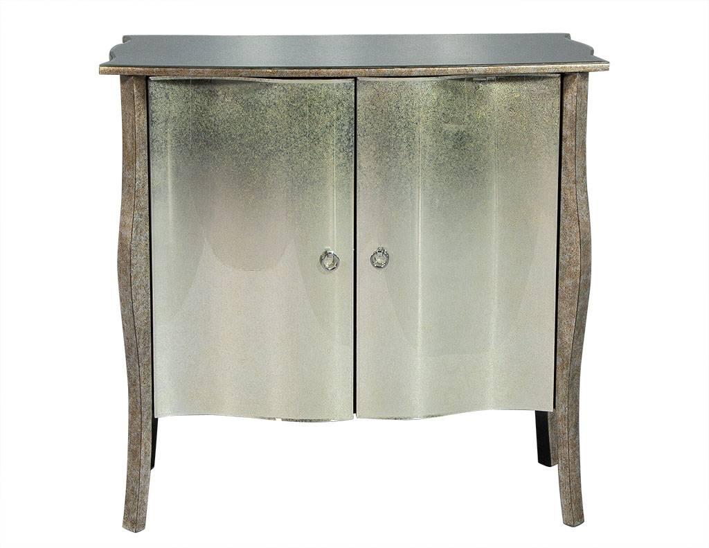 These traditional style cabinets are absolutely striking. They are composed of an antique mirrored, serpentine frame with curved, double doors, accented by a two ring pulls. Floating on four wooden legs in a silver metallic finish, they are a