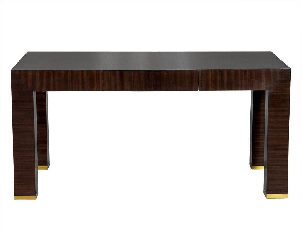 This modern desk is composed of high-gloss, lacquered Macassar ebony and contains a single drawer in the middle. It is also adorned with brass metal trim at the bottom of the four block-shaped, square legs. Simple with a touch of glam, this desk is